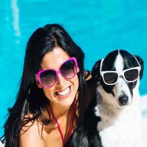 Smiling lady with dog by pool, both are wearing sunglasses